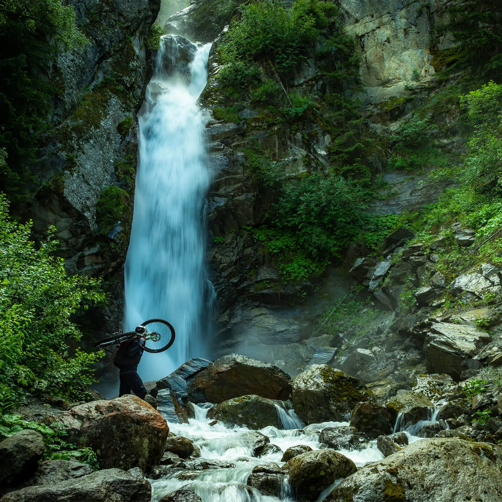 Mountain biker carrying bike near waterfall, representing Mountain Bike Ambassador Management, Mountain Bike Press Camp Planner, and Mountain Bike Event Planner. LOAM.