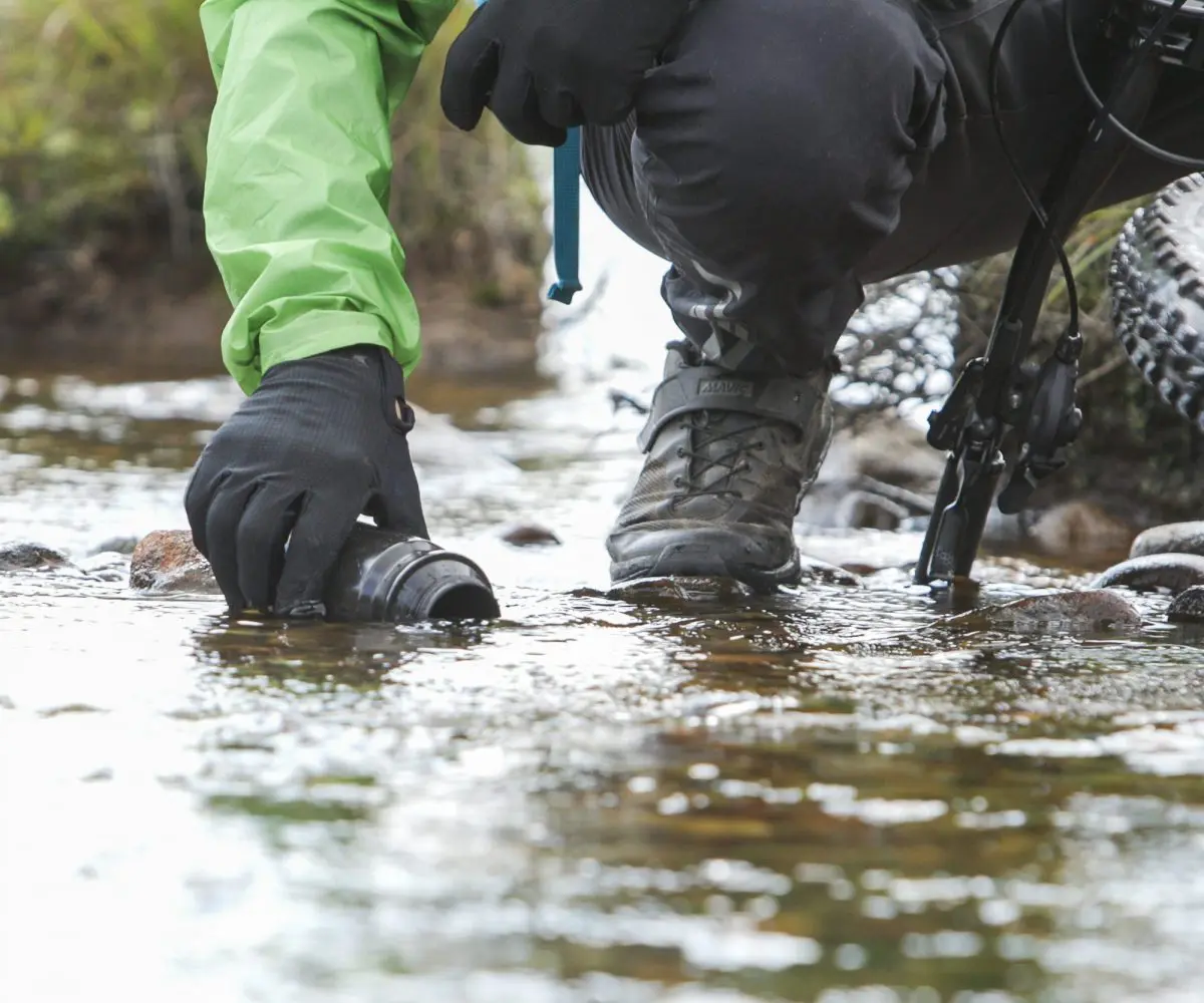 Person in outdoor gear collecting water near mountain bike, ideal for Mountain Bike Ambassador Management and Event Planning content creation strategies. LOAM.