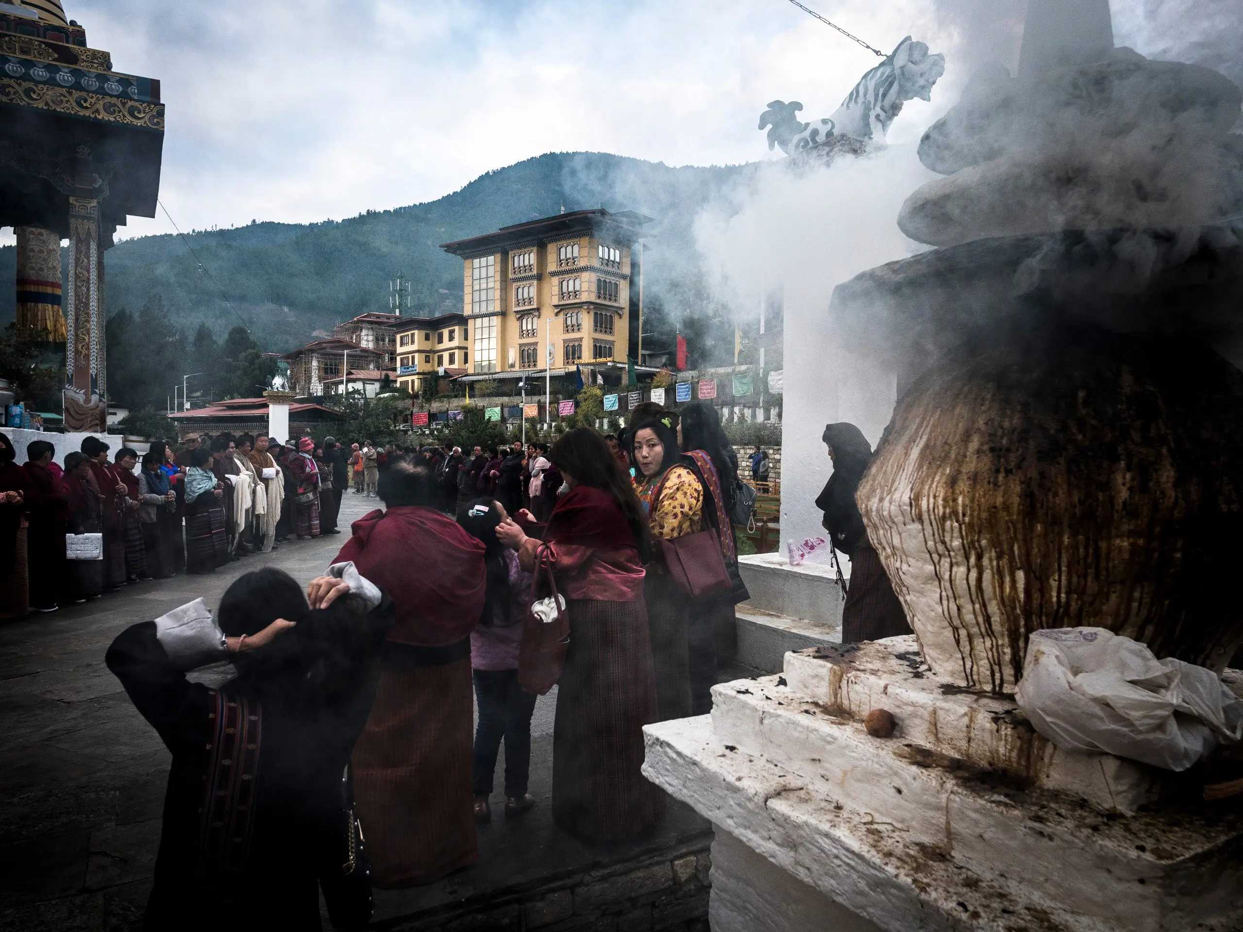 People gather near a smoking incense burner in a Bhutanese courtyard, showcasing local culture. Around the World With LOAM | Euan Wilson’s Global Trail Adventures. LOAM.