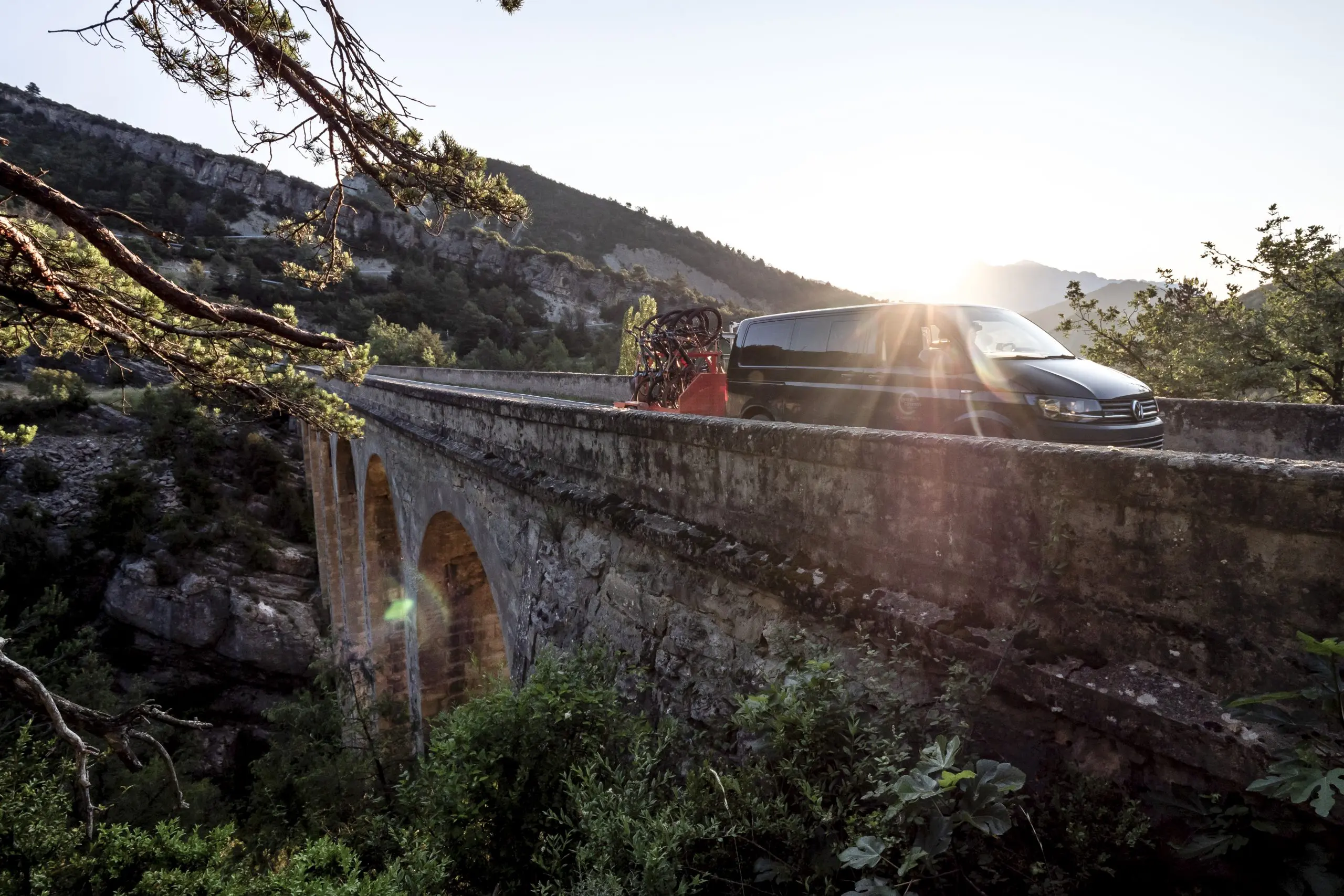 Van transporting mountain bikes on a scenic bridge at sunset. Ideal for Mountain Bike Press Camp and Mountain Bike Event Planner themes. LOAM.