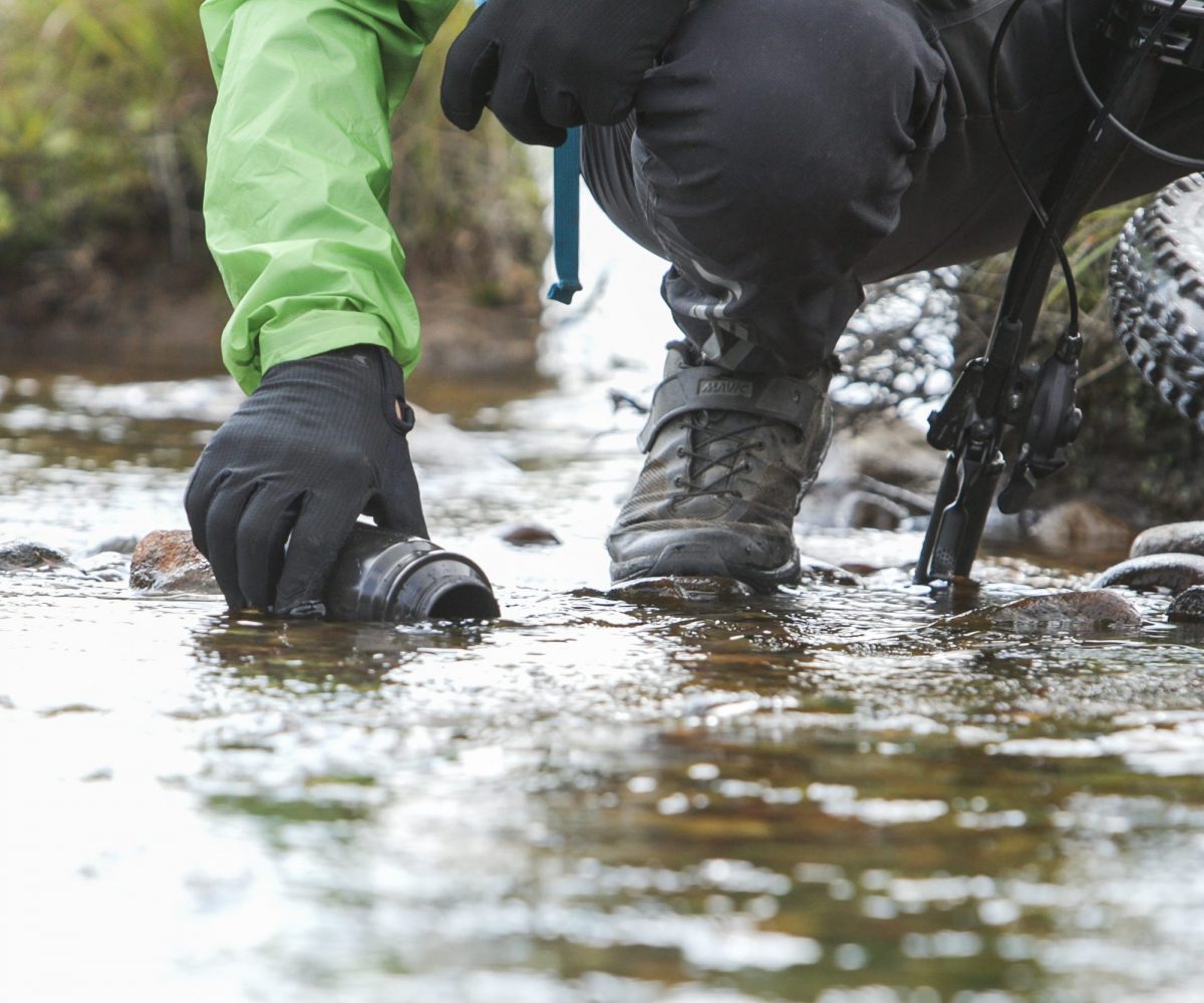 Person in outdoor gear collecting water near mountain bike, ideal for Mountain Bike Ambassador Management and Event Planning content creation strategies. LOAM.